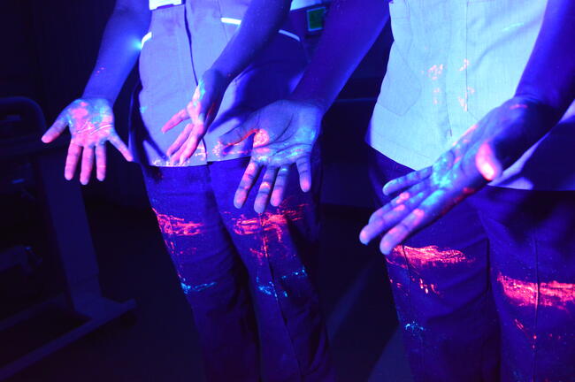 Hands glowing with different coloured mock-pathogens , lit under ultra-violet light.